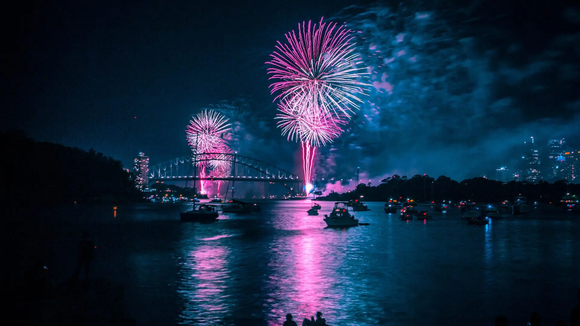imagen de una ciudad por la noche con fuegos artificiales donde se pueden ver embarcaciones en un rio