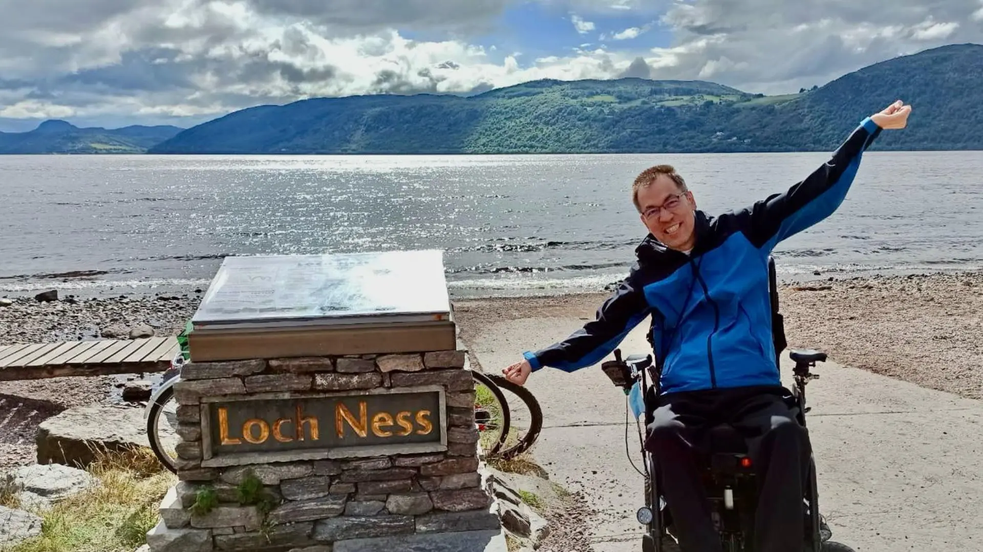 Un usuario de silla de ruedas eléctrica al lado del cartel del Lago Ness, en Escocia