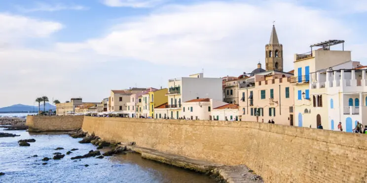 Vistas de la murallas bañadas por el mar que rodean el paseo marítimo de Alghero. Detrás de ellas se aprecian casas de baja altura y la torre de una iglesia. Alghero accesible para personas con discapacidad