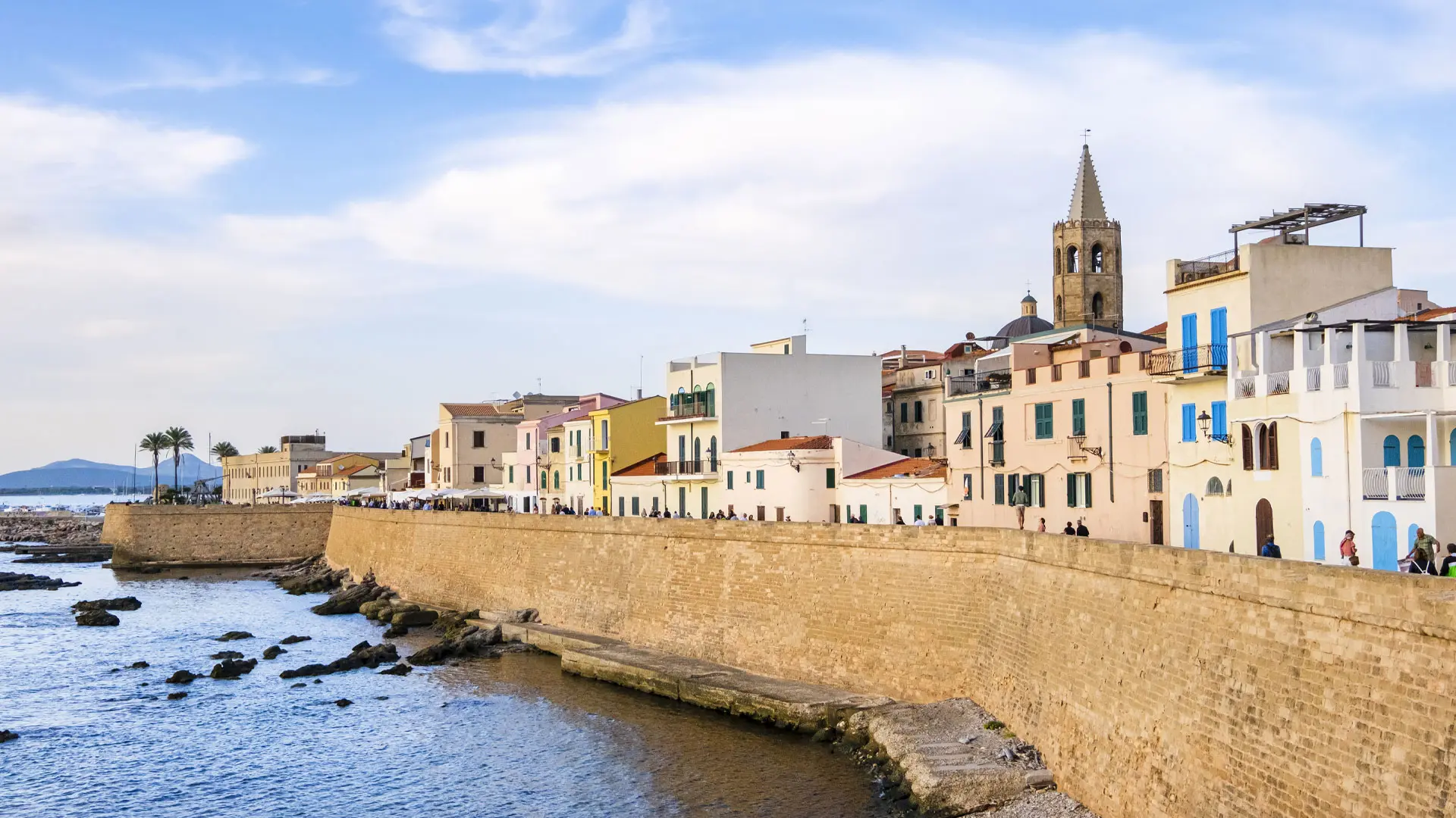 Vistas de la murallas bañadas por el mar que rodean el paseo marítimo de Alghero. Detrás de ellas se aprecian casas de baja altura y la torre de una iglesia. Alghero accesible para personas con discapacidad