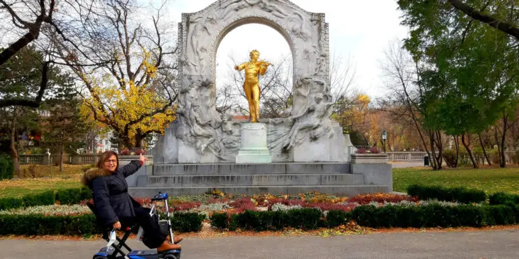 Usuaria de silla de ruedas en al lado izquierdo de la imagen presentando el monumento a Johann Strauss que queda al lado derecho de la imagen. la Viajar a Viena en silla de ruedas.