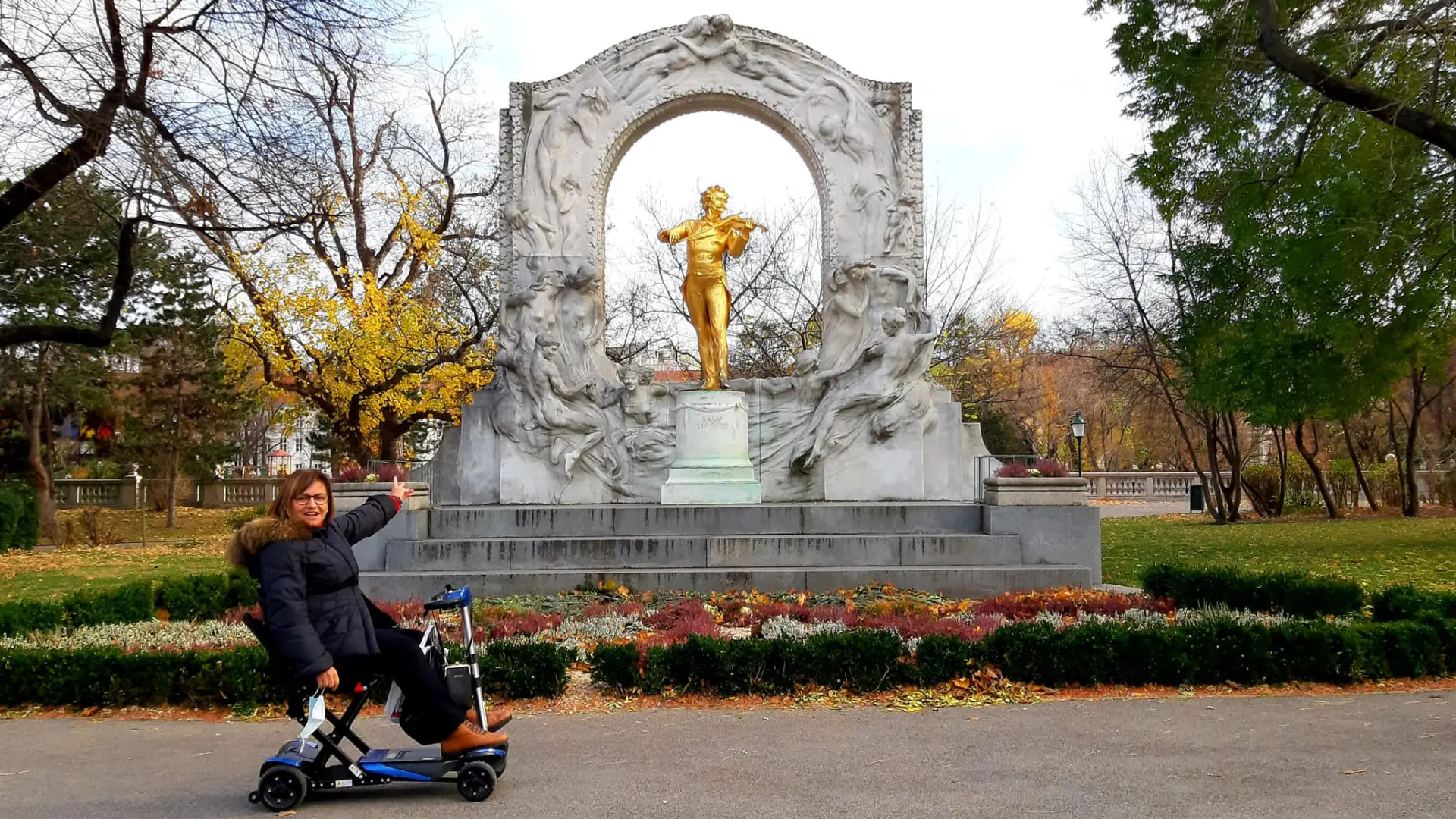 Usuaria de silla de ruedas en al lado izquierdo de la imagen presentando el monumento a Johann Strauss que queda al lado derecho de la imagen. la Viajar a Viena en silla de ruedas.