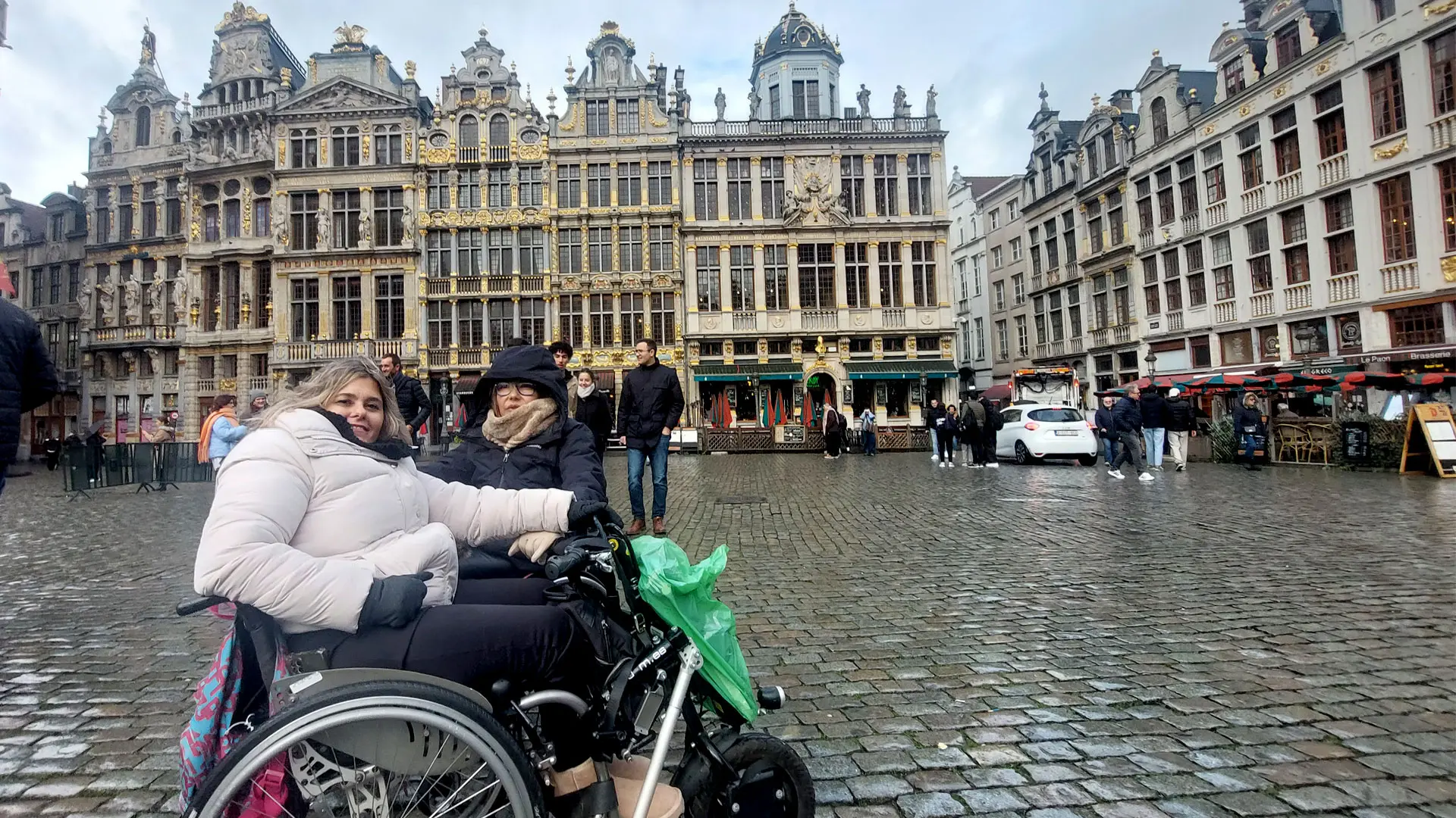 En primera plana dos amigas usuarias de silla de ruedas. Detrás de ellas, de fondo la Grand Place de Bruselas con la arquitectura particular de la zona.