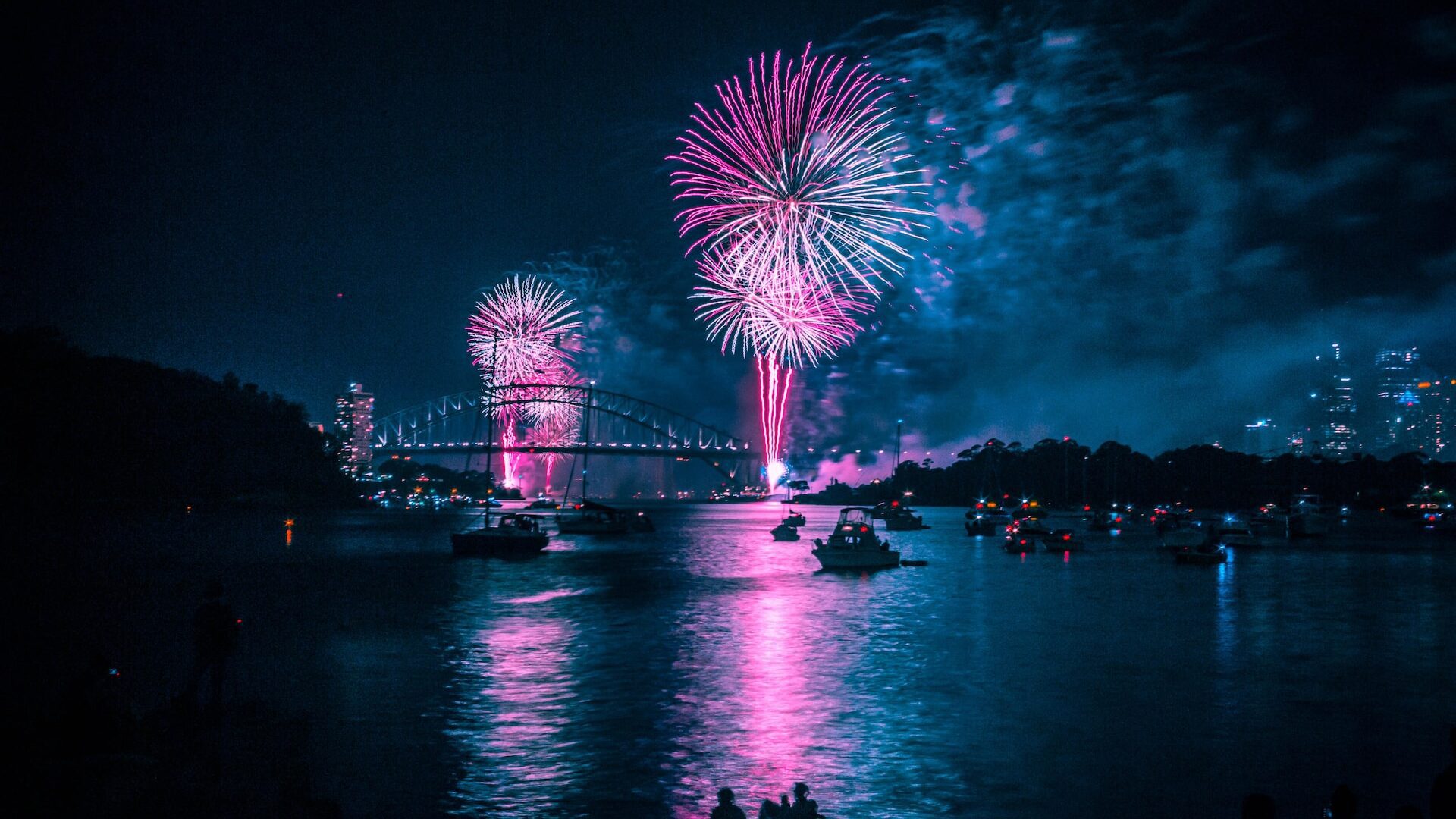 imagen de una ciudad por la noche con fuegos artificiales donde se pueden ver embarcaciones en un rio