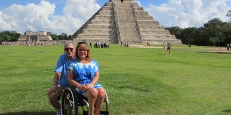 Una pareja viajando por México, ella, usuaria de silla de ruedas, al fondo, la pirámide de Chichén Itzá