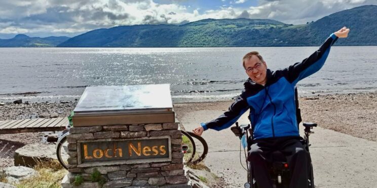 Un usuario de silla de ruedas eléctrica al lado del cartel del Lago Ness, en Escocia