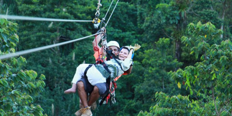 Se ve a dos personas, una de ellas con discapacidad y está realizando una actividad de canopy en Costa Rica siendo asistida por un guía. Están rodeados por las copas de los árboles.