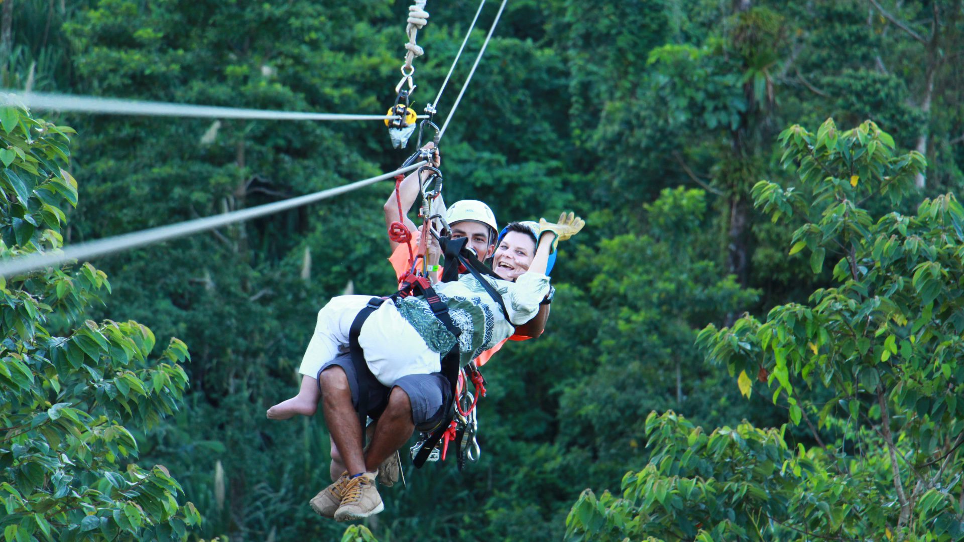 Se ve a dos personas, una de ellas con discapacidad y está realizando una actividad de canopy en Costa Rica siendo asistida por un guía. Están rodeados por las copas de los árboles.