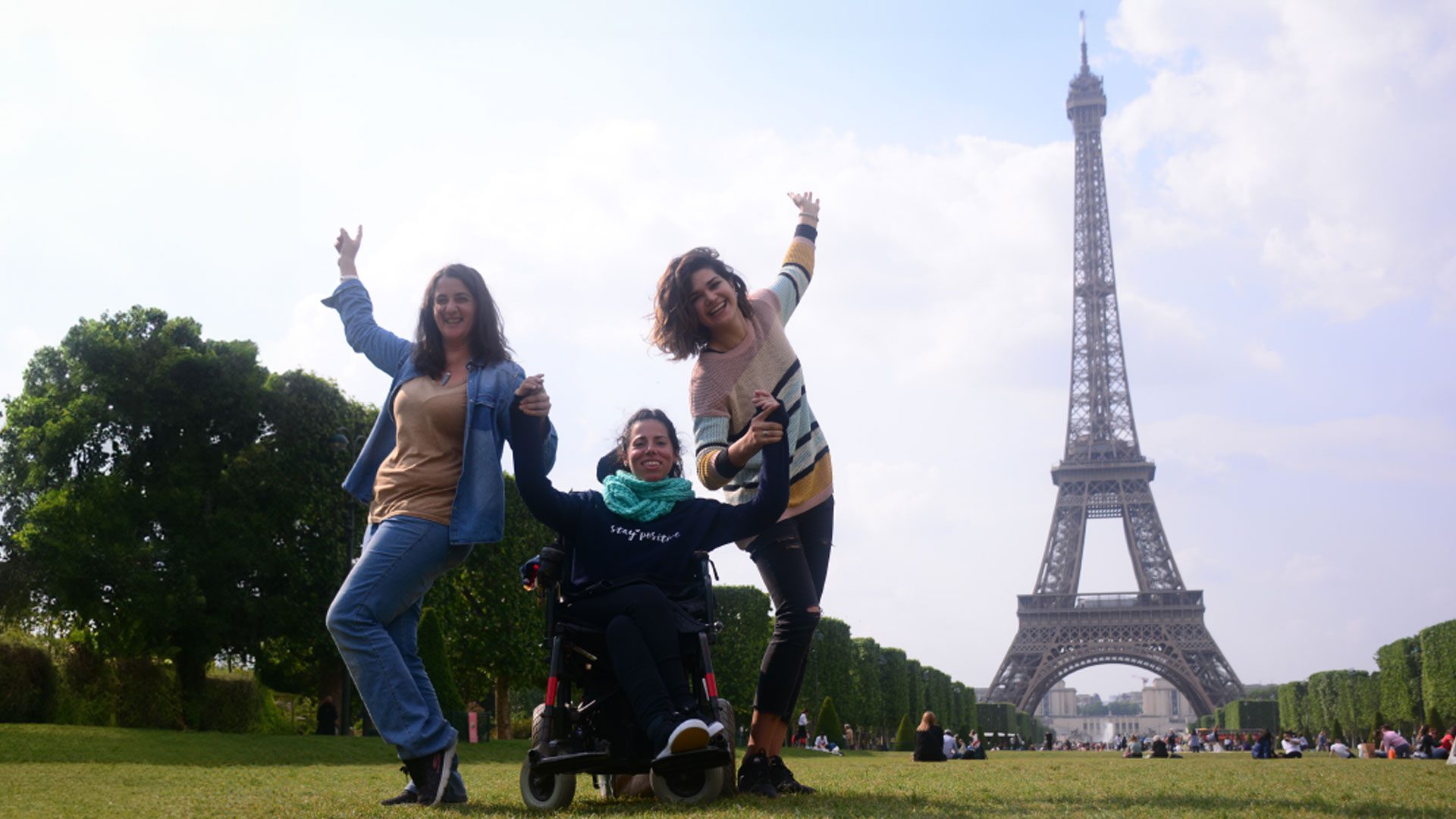 3 amigas, una de ellas usuaria de silla de ruedas, posando para la foto en los jardines con la Torre Eiffel detrás