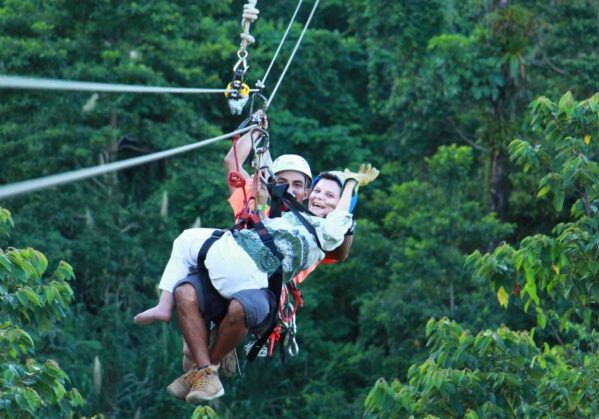 Se ve a dos personas, una de ellas con discapacidad y está realizando una actividad de canopy en Costa Rica siendo asistida por un guía. Están rodeados por las copas de los árboles.