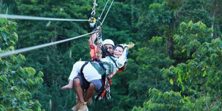 Se ve a dos personas, una de ellas con discapacidad y está realizando una actividad de canopy en Costa Rica siendo asistida por un guía. Están rodeados por las copas de los árboles.