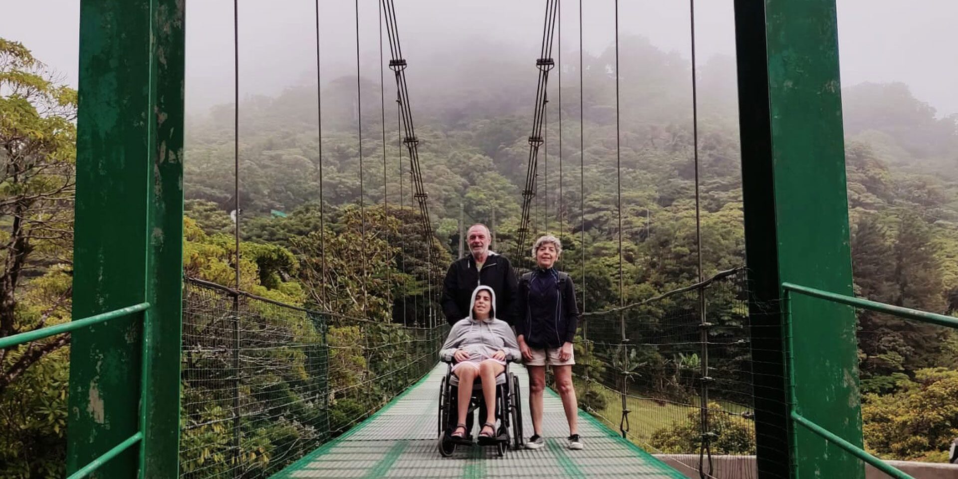 Tres miembros de una familia, la hija usuaria de silla de ruedas, cruzando un puente colgante en Costa Rica. Hay un ambiente nuboso.