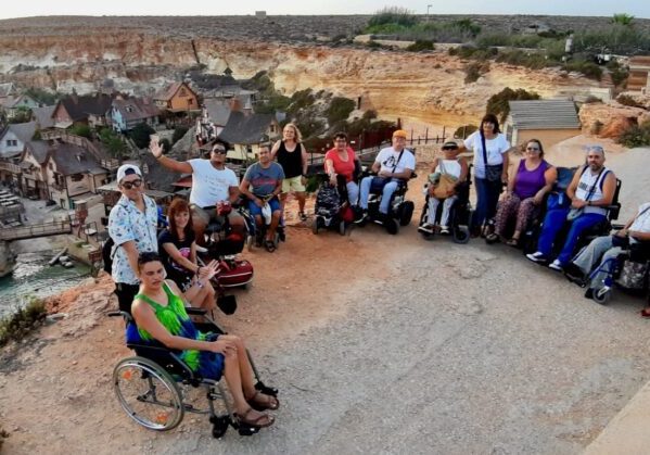 Un grupo de viajeros usuarios de silla de ruedas en un mirador accesible. Al fondo se ve la Villa de Popeye en Malta