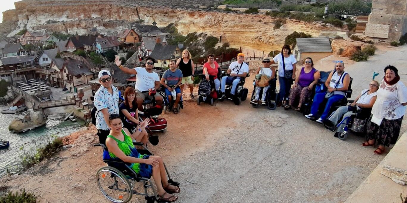 Un grupo de viajeros usuarios de silla de ruedas en un mirador accesible. Al fondo se ve la Villa de Popeye en Malta