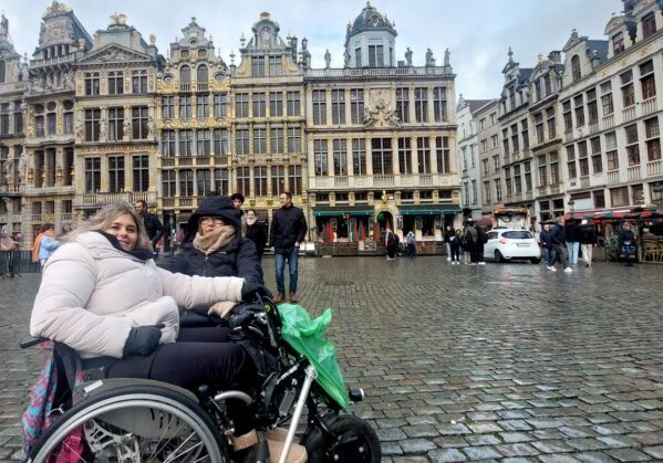 En primera plana dos amigas usuarias de silla de ruedas. Detrás de ellas, de fondo la Grand Place de Bruselas con la arquitectura particular de la zona.