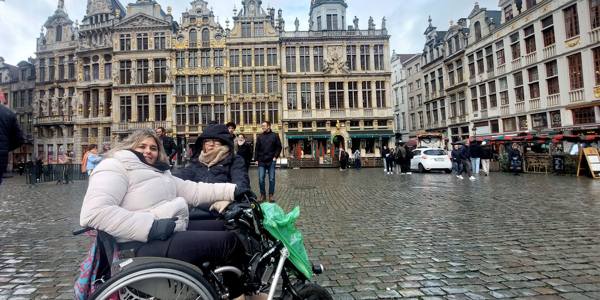 Bruselas en silla de ruedas En primera plana dos amigas usuarias de silla de ruedas. Detrás de ellas, de fondo la Grand Place de Bruselas con la arquitectura particular de la zona.