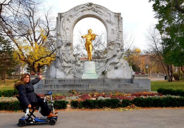 Usuaria de silla de ruedas en al lado izquierdo de la imagen presentando el monumento a Johann Strauss que queda al lado derecho de la imagen. la Viajar a Viena en silla de ruedas.