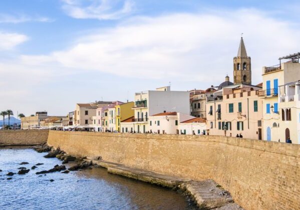 Vistas de la murallas bañadas por el mar que rodean el paseo marítimo de Alghero. Detrás de ellas se aprecian casas de baja altura y la torre de una iglesia. Alghero accesible para personas con discapacidad
