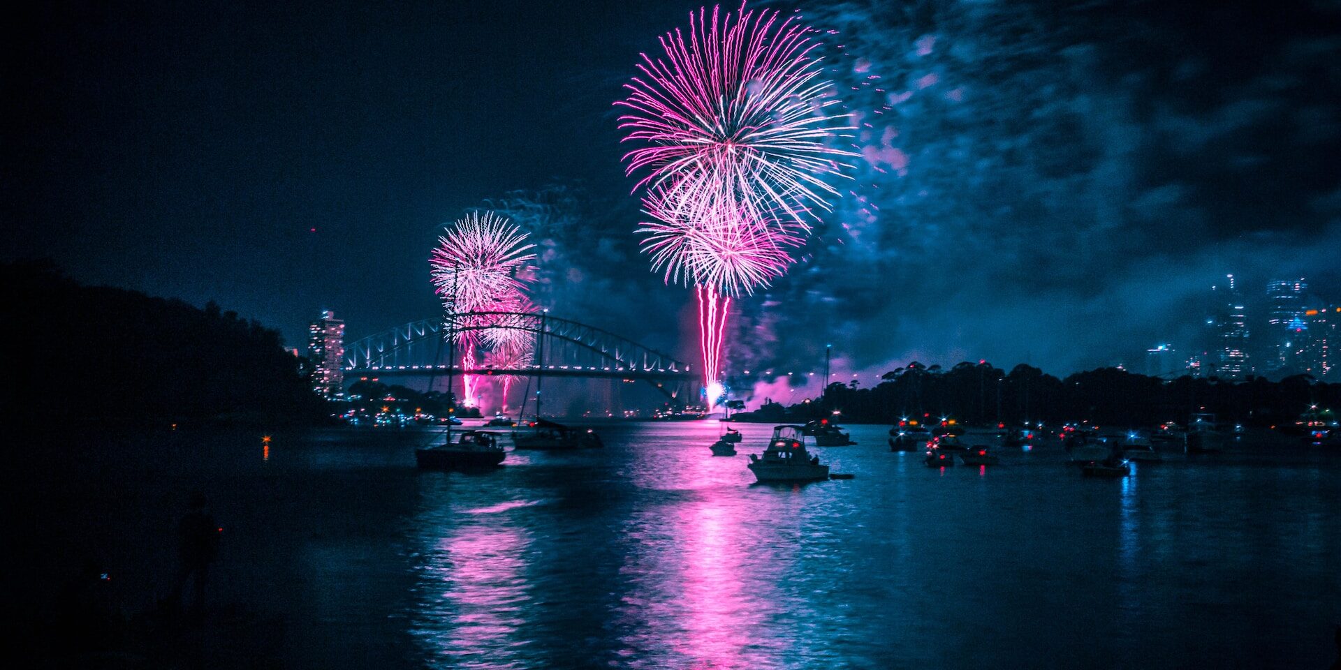 imagen de una ciudad por la noche con fuegos artificiales donde se pueden ver embarcaciones en un rio