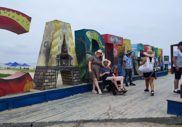 Una pareja, él usuario de silla de ruedas, en un viaje adaptado a Colombia, frente al cartel turístico de Cartagena