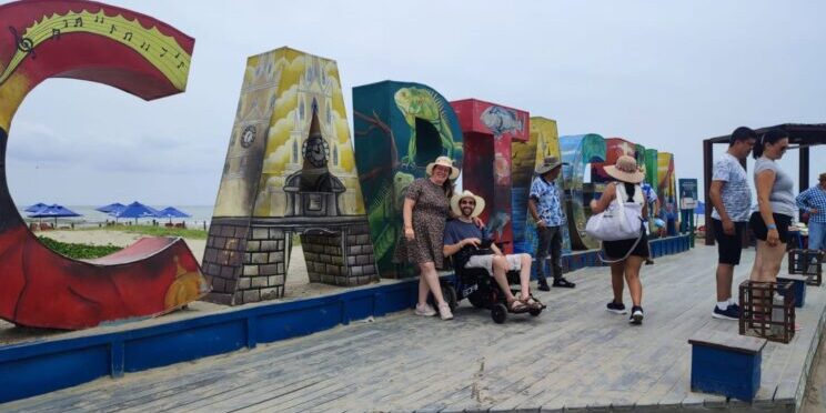 Una pareja, él usuario de silla de ruedas, en un viaje adaptado a Colombia, frente al cartel turístico de Cartagena